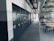 A spacious office or industrial storage space with large metal cabinets lined up along the wall, featuring circular locks. The area also includes modern office chairs and workstations with orange accents. Overhead, there is an array of exposed pipes and ventilation ducts.