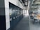 A spacious office or industrial storage space with large metal cabinets lined up along the wall, featuring circular locks. The area also includes modern office chairs and workstations with orange accents. Overhead, there is an array of exposed pipes and ventilation ducts.