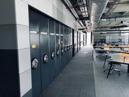 A spacious office or industrial storage space with large metal cabinets lined up along the wall, featuring circular locks. The area also includes modern office chairs and workstations with orange accents. Overhead, there is an array of exposed pipes and ventilation ducts.