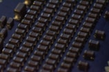 Close-up view of a circuit board featuring rows of small, rectangular electronic components that appear to be capacitors or resistors. The components are evenly arranged in parallel lines across a dark surface, likely the base of the circuit board. The lighting creates a slightly reflective surface, emphasizing the precise layout.