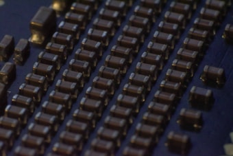 Close-up view of a circuit board featuring rows of small, rectangular electronic components that appear to be capacitors or resistors. The components are evenly arranged in parallel lines across a dark surface, likely the base of the circuit board. The lighting creates a slightly reflective surface, emphasizing the precise layout.