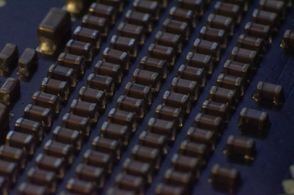 Close-up view of a circuit board featuring rows of small, rectangular electronic components that appear to be capacitors or resistors. The components are evenly arranged in parallel lines across a dark surface, likely the base of the circuit board. The lighting creates a slightly reflective surface, emphasizing the precise layout.
