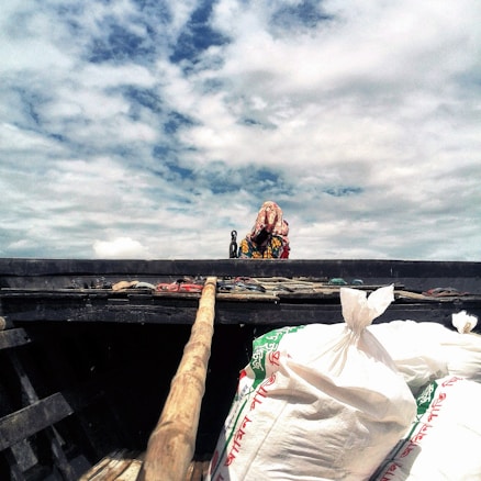 A person wrapped in colorful fabric is standing on a platform, seemingly on a boat or dock. The sky is mostly cloudy with patches of blue. Large sacks, possibly of goods or supplies, are visible in the foreground, along with a wooden pole extending towards the top of the image.