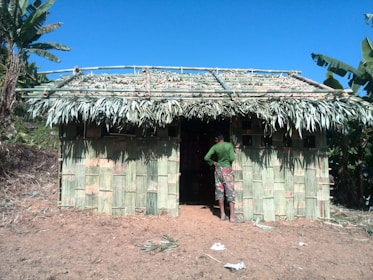 A small, rustic hut constructed from green bamboo and palm leaves stands on a patch of bare earth. The roof is made of thick layers of palm fronds, while the walls are woven bamboo. A person dressed in a green long-sleeve shirt and patterned pants faces the hut, suggesting a rural or agricultural setting. Surrounding the hut are leafy banana trees under a clear blue sky.