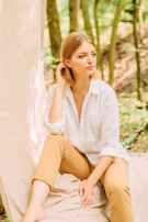 A person with blonde hair is seated outdoors. They are wearing a white long-sleeved shirt with the sleeves rolled up and tan pants. The individual appears to be thoughtful or contemplative, with one hand touching their hair and the other resting on their knee. The backdrop consists of a soft fabric draped vertically next to the person, with green foliage and tree trunks in the background, suggesting a natural, forest setting.