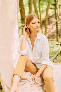 A person with blonde hair is seated outdoors. They are wearing a white long-sleeved shirt with the sleeves rolled up and tan pants. The individual appears to be thoughtful or contemplative, with one hand touching their hair and the other resting on their knee. The backdrop consists of a soft fabric draped vertically next to the person, with green foliage and tree trunks in the background, suggesting a natural, forest setting.