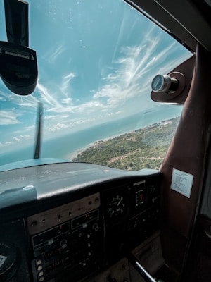 A view from the cockpit of a small aircraft, showcasing the dashboard with various gauges and controls. Through the windshield, a scenic landscape with a coastline and a stretch of blue ocean can be seen under a sky filled with wispy clouds.
