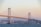 A large suspension bridge connects two landmasses over a body of water. Tall towers and cables support the structure, which is surrounded by an urban landscape featuring numerous buildings. The sky is clear with a warm tone, suggesting early morning or late afternoon lighting.