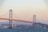 A large suspension bridge connects two landmasses over a body of water. Tall towers and cables support the structure, which is surrounded by an urban landscape featuring numerous buildings. The sky is clear with a warm tone, suggesting early morning or late afternoon lighting.