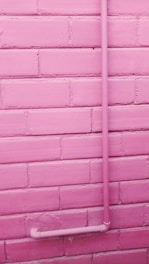 A pink-painted brick wall with a pink pipe attached running vertically up and then turning horizontally near the bottom.
