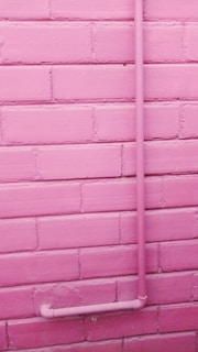 A pink-painted brick wall with a pink pipe attached running vertically up and then turning horizontally near the bottom.