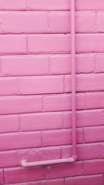 A pink-painted brick wall with a pink pipe attached running vertically up and then turning horizontally near the bottom.
