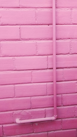 A pink-painted brick wall with a pink pipe attached running vertically up and then turning horizontally near the bottom.