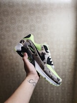 A hand with a tattoo holding a camouflage-patterned sneaker featuring neon green accents and a black swoosh logo. The shoe is an Air Max model, positioned against a decorative beige wallpaper.