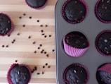 Several chocolate muffins are displayed on a wooden surface and inside a muffin tray. The muffins are topped with chocolate chips and are placed in pink wrappers. Some chocolate chips are scattered on the wooden surface.