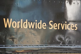 Golden text displaying 'Worldwide Services' is set against a dark, reflective surface, likely part of a vehicle or metallic structure, featuring rivets and a slightly worn texture.