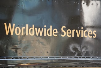 Golden text displaying 'Worldwide Services' is set against a dark, reflective surface, likely part of a vehicle or metallic structure, featuring rivets and a slightly worn texture.