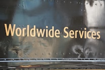 Golden text displaying 'Worldwide Services' is set against a dark, reflective surface, likely part of a vehicle or metallic structure, featuring rivets and a slightly worn texture.