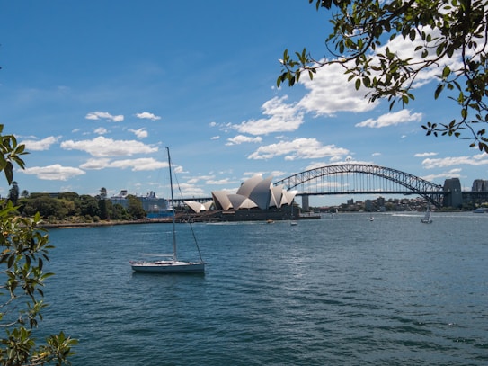 A scenic view of a large, iconic opera house near the edge of a blue body of water with a sailboat in the foreground. Beyond the opera house, an arched steel bridge extends across the water. The sky is clear with scattered fluffy clouds, and leafy branches frame parts of the image.