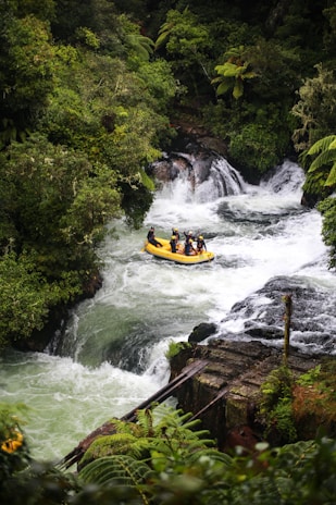A group of people is navigating a rapid section of a river on a yellow inflatable raft surrounded by lush green vegetation and ferns. The water is white and turbulent as it flows over rocks and small waterfalls.