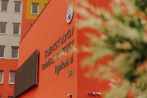 A vibrant orange building facade with text in a foreign language. The structure has multiple windows and a security camera mounted on the wall. A circular emblem is displayed on the upper part of the building. The top left corner shows part of another building with windows. A blurred plant is partially visible in the foreground.