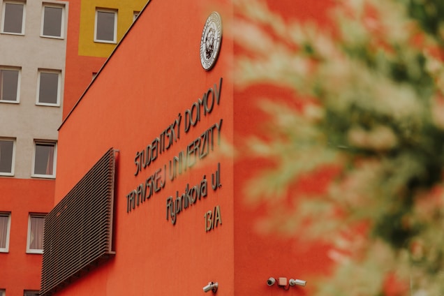 A vibrant orange building facade with text in a foreign language. The structure has multiple windows and a security camera mounted on the wall. A circular emblem is displayed on the upper part of the building. The top left corner shows part of another building with windows. A blurred plant is partially visible in the foreground.