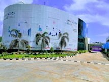 A modern building with a curved facade featuring stylized illustrations and text promoting innovation. Tall palm trees line the front, and a well-maintained garden enhances the entrance area. The sky is clear with few clouds, suggesting a bright day.