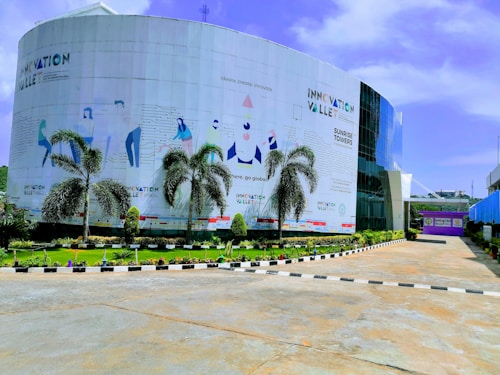 A modern building with a curved facade featuring stylized illustrations and text promoting innovation. Tall palm trees line the front, and a well-maintained garden enhances the entrance area. The sky is clear with few clouds, suggesting a bright day.