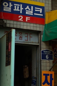 A storefront entrance with signage in Korean, displaying a blue and red sign indicating '2F'. Adjacent is a tiled wall with a street sign reading 'Eulji-ro 16-gil'. The entrance appears dimly lit, with an open door and some posters on the wall. There is a portion of a yellow and green awning visible on the right side.