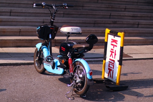 A blue electric scooter is parked next to a yellow and black sign with red Korean characters. The scene is set on a paved area near steps, suggesting an urban environment. The scooter has a brown seat and is positioned in the sunlight.