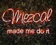 A vibrant neon sign with the words 'Mezcal made me do it' is surrounded by lush green foliage and plants. The neon lights are in an eye-catching orange-red color.