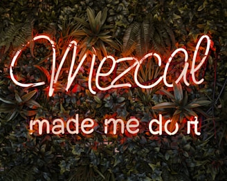 A vibrant neon sign with the words 'Mezcal made me do it' is surrounded by lush green foliage and plants. The neon lights are in an eye-catching orange-red color.