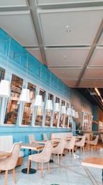 Bright and modern cafe with blue walls, white ceilings with panel detailing, and rows of white pendant lights. The seating consists of beige cushioned chairs and tables with light wood surfaces. There are large windows with reflections indicating an interior setting. The overall style is contemporary and clean with an inviting ambiance.