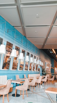 Bright and modern cafe with blue walls, white ceilings with panel detailing, and rows of white pendant lights. The seating consists of beige cushioned chairs and tables with light wood surfaces. There are large windows with reflections indicating an interior setting. The overall style is contemporary and clean with an inviting ambiance.