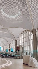 A spacious interior with a modern, curved ceiling made of metal slats and large windows allowing natural light to flood in. The area features digital screens, glass barriers, and a sleek, minimalist design.