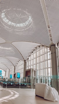 A spacious interior with a modern, curved ceiling made of metal slats and large windows allowing natural light to flood in. The area features digital screens, glass barriers, and a sleek, minimalist design.