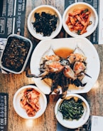 A variety of Korean dishes are artfully arranged on a table. At the center of the spread, there is a large plate with marinated crab, surrounded by small dishes containing different side dishes including kimchi, spicy radish salad, seaweed, and greens. The table has a textured surface and some menu text in Korean is visible.