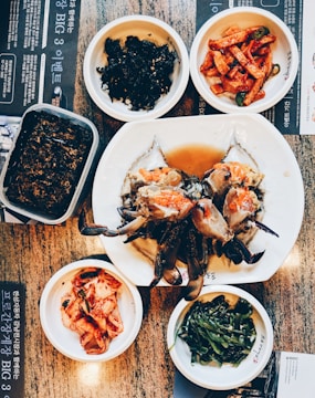 A variety of Korean dishes are artfully arranged on a table. At the center of the spread, there is a large plate with marinated crab, surrounded by small dishes containing different side dishes including kimchi, spicy radish salad, seaweed, and greens. The table has a textured surface and some menu text in Korean is visible.