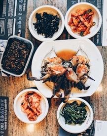 A variety of Korean dishes are artfully arranged on a table. At the center of the spread, there is a large plate with marinated crab, surrounded by small dishes containing different side dishes including kimchi, spicy radish salad, seaweed, and greens. The table has a textured surface and some menu text in Korean is visible.