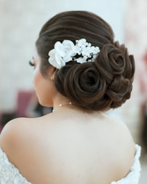 A woman is seen from the back showcasing an intricate hairstyle featuring large, smooth curls pinned into a low bun. Adorned with a white floral hairpiece, the style is elegant and sophisticated. The exposed shoulders suggest formal or bridal attire.