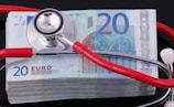 A stethoscope with red tubing rests on a stack of 20 euro banknotes, with the stethoscope's head placed centrally on the top banknote. The banknotes are neatly stacked on a reflective black surface, creating a sharp contrast.