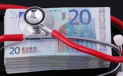 A stethoscope with red tubing rests on a stack of 20 euro banknotes, with the stethoscope's head placed centrally on the top banknote. The banknotes are neatly stacked on a reflective black surface, creating a sharp contrast.