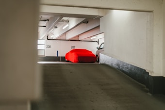 A car covered with a bright red fabric sits parked in a dimly lit parking garage. The numbers '207' and '208' are visible on the walls, indicating parking spaces. The garage has a concrete structure with a sloping floor and fluorescent lights on the ceiling.