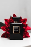 A sleek black perfume bottle labeled Coco Noir Chanel Paris is positioned against a backdrop of vibrant red flowers. The bottle's simplistic design and gold accents exude luxury and elegance. The contrast between the dark bottle and the bright flowers creates a striking visual appeal.