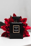 A sleek black perfume bottle labeled Coco Noir Chanel Paris is positioned against a backdrop of vibrant red flowers. The bottle's simplistic design and gold accents exude luxury and elegance. The contrast between the dark bottle and the bright flowers creates a striking visual appeal.
