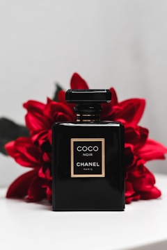 A sleek black perfume bottle labeled Coco Noir Chanel Paris is positioned against a backdrop of vibrant red flowers. The bottle's simplistic design and gold accents exude luxury and elegance. The contrast between the dark bottle and the bright flowers creates a striking visual appeal.