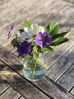 A glass table vase filled with fresh wildflowers on a sunlit wooden table.