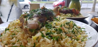 A plate of rice garnished with fresh herbs and topped with a cooked piece of meat, likely chicken or lamb. The rice appears to contain small pine nuts and is served in a bright and airy setting with a window in the background showing a blurred view of the street and some parked cars. A glass pitcher of water with a slice of lemon and mint leaves is placed alongside, adding a refreshing touch.