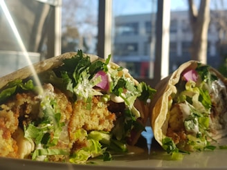 A close-up of a wrap filled with crispy falafel, fresh greens, purple onions, and a drizzle of creamy sauce. The wrap is cut in half, displaying the colorful and textured filling. Sunlight streams through a nearby window, casting a warm glow over the plate.