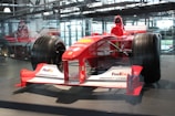 A red Formula 1 race car displayed in an indoor setting, featuring logos from various sponsors like FedEx, Bridgestone, and Magneti Marelli. The car is surrounded by a glass enclosure, with natural light filtering through the large windows in the background. The sleek design and large tires of the vehicle are prominently visible.